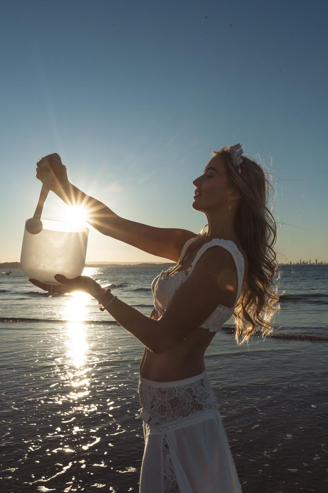 Bryony lifting a crystal singing bowl into the setting sun at the water's edge